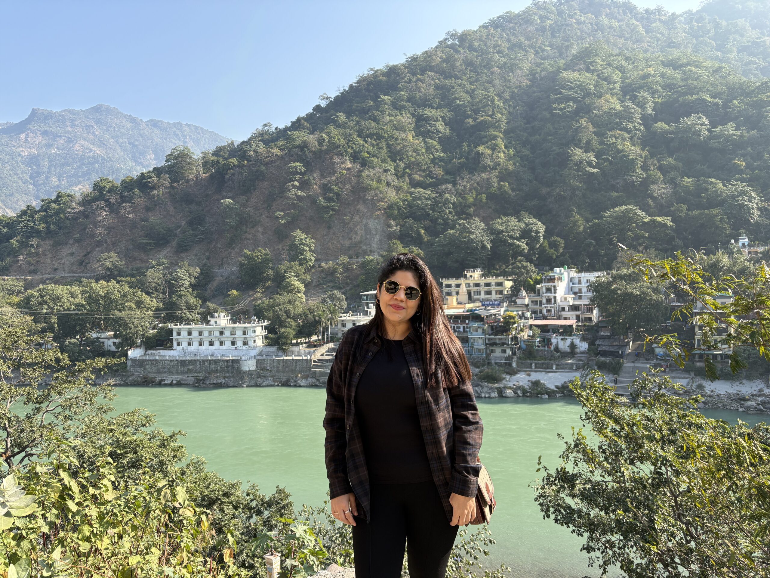 Solo woman traveller enjoying her first weekend trip to Rishikesh with Jugni Travel, standing by the Ganga River with lush green hills in the background.